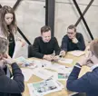 Vijf collega’s rond een houten tafel bespreken drukwerk en foto’s in een moderne vergaderruimte.