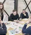 Vijf collega’s rond een houten tafel bespreken drukwerk en foto’s in een moderne vergaderruimte.