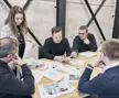 Vijf collega’s rond een houten tafel bespreken drukwerk en foto’s in een moderne vergaderruimte.